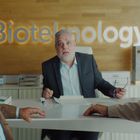 Text: "Biotelnology." Drei Männer schauen überrascht, zwei im Vordergrund, einer am Schreibtisch sitzend im Anzug.