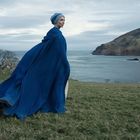 Eine Person in einem langen, blauen Umhang steht auf einer grünen Wiese am Meer mit Hügeln im Hintergrund.