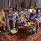 Große Gruppe von Menschen sitzt auf einem Sofa, schaut konzentriert und glücklich. Laptop, Snacks, Getränke und Popcorn sind vorhanden.
