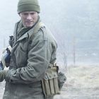 Ein Mann in Militärkleidung und Mütze hält eine Waffe, während er in einer nebligen, bewaldeten Umgebung steht.