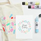 "Happy Birthday" in eleganter Schrift auf Karte, umgeben von bunten Punkten und Formen, liegt neben Stofftasche und 3D-Stiften.