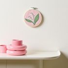 Ein runder Stickrahmen mit Maiglöckchen-Motiv an weißer Wand. Daneben rosafarbene Keramikvasen auf einem Regal.