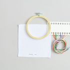 Stickrahmen, Stoffquadrat, bunte Garnfäden durch ein Lochbrett gefädelt auf grauem Hintergrund.