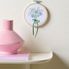 Eine Vase steht auf einem Regal, daneben hängt ein rundes Stickbild mit bunten Blumen und grünem Band.