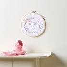 „WELCOME HOME“ in einem Stickrahmen mit Blumenkranz; darunter rosa Stapelbecher auf einem Tisch.