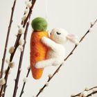 Ein Filzhase umarmt eine große, orange Karotte, die an blühenden Zweigen hängt.