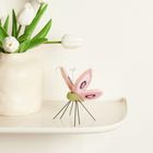 Weiße Tulpen in einer Keramikvase, daneben eine stilisierte Schmetterlingsfigur mit rosa Flügeln auf einem Regal.