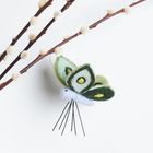 Zweig mit flauschigen Knospen im Vordergrund, daran hängt ein Filzschmetterling mit grün-gelbem Muster.