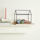 Ein schwarzes Drahtmodell eines Hauses steht auf einem Regal neben einem rosa gedrehten Kerzenständer. Darin sind bunte Eier.