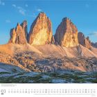 03. März. Drei Zinnen, Dolomiten. Felsformationen unter blauem Himmel bei Sonnenaufgang, im Vordergrund grüne Wiesen.