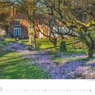 2, 2027. Ein Garten mit blühenden lila Krokussen, Bäumen und einem roten Backsteinhaus im Hintergrund.