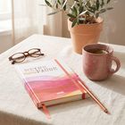 „MEINE menoPAUSE“ auf einem Buch, Brille, Tasse Tee, Topfpflanze, Stift auf einem hellen Tischtuch.