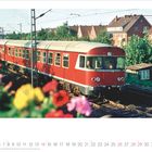 Zahlen 1 bis 31 unten; März 2027. Ein roter Zug fährt auf Gleisen, im Vordergrund unscharfe Blumen, im Hintergrund Häuser.