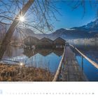 Bootshäuser am Kochelsee, Bayern, Deutschland. Steg führt über Wasser. Klare Bergkulisse, strahlende Sonne hinter Bäumen.
