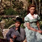 Zwei Personen in traditioneller Kleidung sitzen im Freien. Eine Frau trägt ein grünes Kleid, ein Mann einen schwarzen Hut.