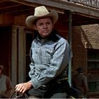 Ein Mann in Cowboy-Kleidung und Hut sitzt auf einem Pferd vor einem Gebäude in einer Western-Kulisse.