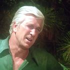 Ein älterer Mann mit weißem Haar in einem grünen Hemd steht im Sonnenlicht vor grünen Nadelzweigen im Hintergrund.