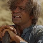 Ich kann keine Texte lesen oder erkennen. Ein Mann mit zotteligem Haar hält einen Stock in einem sandigen Hintergrund.