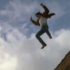 Ein Mensch springt in der Luft, hält eine Waffe. Im Hintergrund sind Wolken und ein Gebäude zu sehen.