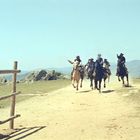 Vier Reiter galoppieren über einen staubigen Weg, umgeben von felsiger Landschaft und Bergen im Hintergrund. Links ein Holzzaun.