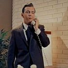 Ein Mann im Anzug telefoniert mit einem alten Telefon. Im Hintergrund sieht man eine Zimmerpflanze und eine helle Backsteinwand.