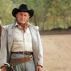 Ein älterer Cowboy in Westernkleidung steht vor einem Planwagen. Im Hintergrund sind Bäume zu sehen.