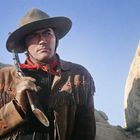Ein Cowboy mit Hut und Gewehr steht in einer felsigen Landschaft. Neben ihm eine Person in einer Decke gehüllt.