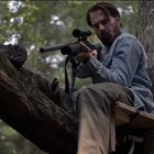 Ein Mann in Jeansjacke sitzt auf einem Baum und zielt mit einem Gewehr. Waldumgebung.