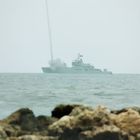 Kein Text im Bild. Ein großes Kriegsschiff feuert etwas ab, während es auf dem Meer fährt. Steine im Vordergrund.