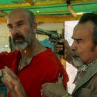 Zwei Männer, der eine bedroht den anderen mit einer Waffe. Der Bedrohte trägt ein rotes Hemd und hebt die Hände.