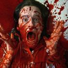 Eine Person schreit erschrocken, bedeckt von einer roten Flüssigkeit, die überall im Raum verteilt ist.