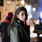 Ich kann den Text auf dem Bild nicht erkennen. Eine Frau blickt ernst in die Kamera, im Hintergrund unscharfe Stadtlichter.