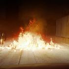 Ein brennender Müllhaufen in einer Gasse, umgeben von Wänden. Links stehen Mülltonnen.