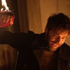 Ein Mann mit Bart hält eine brennende rote Hand. Er wirkt aufgeregt. Im Hintergrund ist warmes Licht zu sehen.