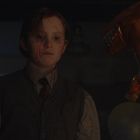 Ein Kind mit neutralem Ausdruck, Tröpfchen auf seinem Gesicht, neben einem orangenen Dinosaurierballon und anderen Luftballons.