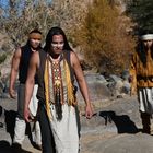 Drei Männer in traditioneller Kleidung und Gesichtsbemalung stehen in einer felsigen Landschaft mit Bäumen im Hintergrund.