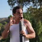 Ein Mann in einem weißen Tanktop pustet in ein Rohr, sein Gesicht ist verzerrt, seine Hand zeigt ein "Okay"-Zeichen.