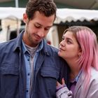 Zwei Personen stehen eng beisammen. Eine Person mit pinken Haaren blickt den anderen liebevoll an. Im Hintergrund Marktstände.