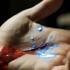 Eine Hand, die blutrot und leuchtend blau gefärbt ist; blaue Tropfen fallen hinein.