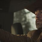 Ein bärtiger Mann in einem Auto, trägt einen Cowboyhut und schaut konzentriert nach vorne, Hände am Lenkrad.