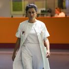 Keine sichtbaren Texte. Eine Frau in weißem Arztkittel geht ernst durch einen Korridor in einem Krankenhaus. Im Hintergrund ist eine Rezeption zu sehen.