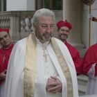 Ein Priester in weißer Robe mit goldener Stola steht vor mehreren Kardinälen in roten Gewändern. Im Hintergrund ein Schweizer Gardist.
