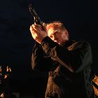 Ein Mann in dunkler Kleidung zielt mit einer Waffe nach oben, in einer nächtlichen Szenerie mit gedämpftem Licht.