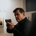 Ein Mann im schwarzen Hemd hält eine Pistole, schaut konzentriert; im Hintergrund ein unscharfes Bild an der Wand.