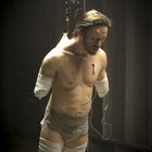 Ein Mann ohne Gliedmaßen, hängt mit Verbänden, ein Infusionsbeutel über ihm, erscheint nachdenklich und erschöpft.