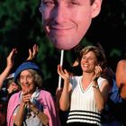 Eine Menschenmenge jubelt; eine Frau hält lächelnd ein großes Gesichtsporträt hoch. Eine ältere Dame klatscht daneben.