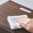 Eine Hand wischt mit einem weißen Tuch Wassertropfen von einer Holzoberfläche mit Griffloch.