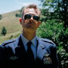 U.S. ist zu lesen. Ein Mann in Uniform mit Sonnenbrille steht vor einer grünen, hügeligen Landschaft.