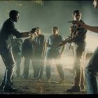 Männer stehen in Konfrontation, einer hält eine Waffe, Beleuchtung von einem Auto im Hintergrund, angespannte Stimmung.