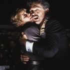 Ein Mann umarmt eine Frau ängstlich. Beide wirken erschrocken. Die Szene ist dunkel, im Hintergrund leuchtet eine Stadt.